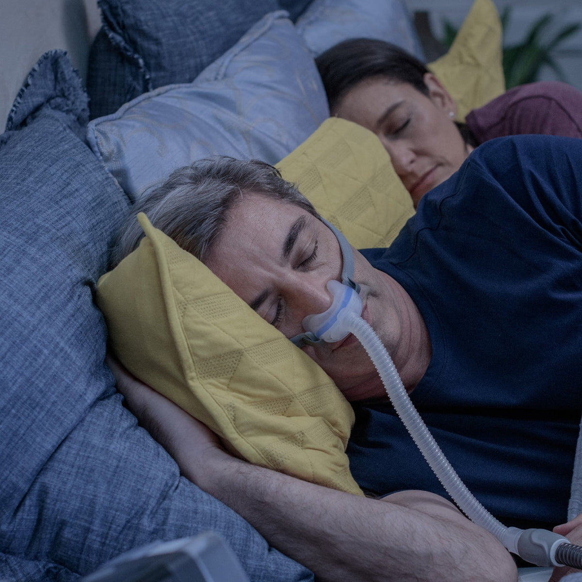 Man asleep on his side wearing the AirFit N30, the cradle cushion under his nose, partner sleeping beside him