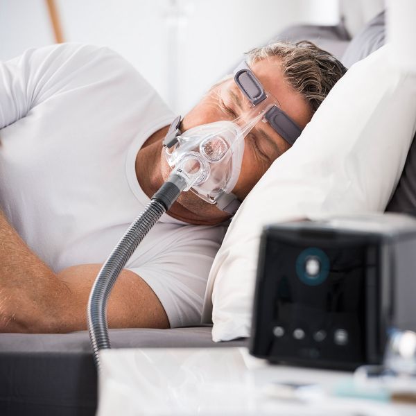 A man sleeping peacefully while wearing a full-face CPAP mask connected to a ResMed AirSense 10 machine on his nightstand.