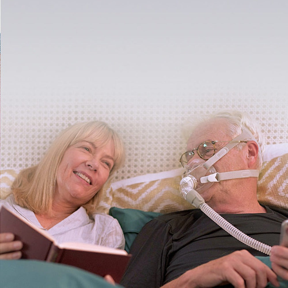Man wearing an AirFit F30 mask while reading in bed beside his partner, the tube running in front
