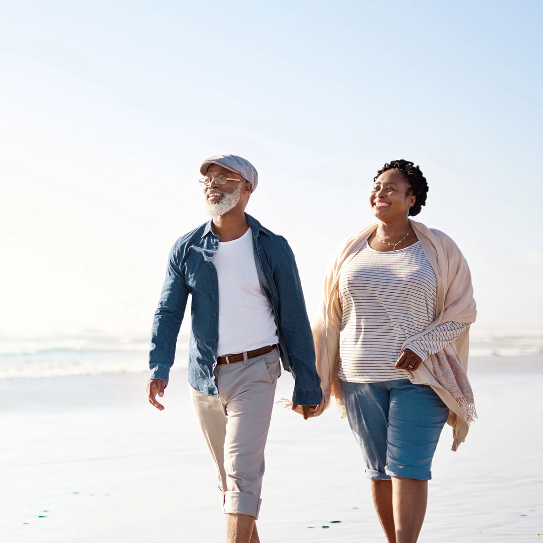 An older couple walking barefoot along a sunny beach, smiling and holding hands