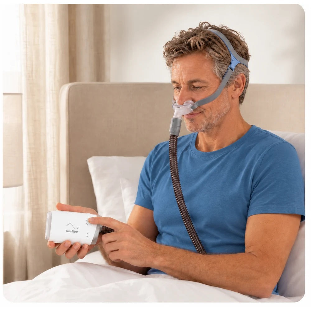 Photograph of a man sitting up in bed wearing a nasal pillows mask while he holds the AirMini AutoSet