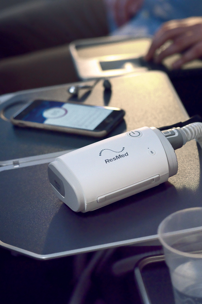 Photograph of the AirMini AutoSet on an airplane tray table beside a phone, with its tube attached
