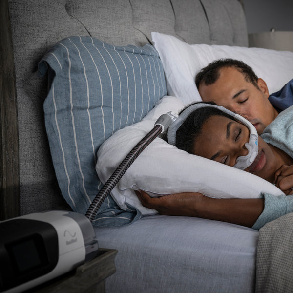 Couple asleep in bed, one wearing a nasal mask connected by heated tubing to an AirSense 11 on the nightstand