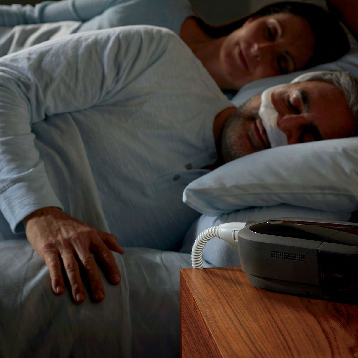 A couple asleep in bed, the man in a nasal CPAP mask, with the DreamStation 2 on the bedside table