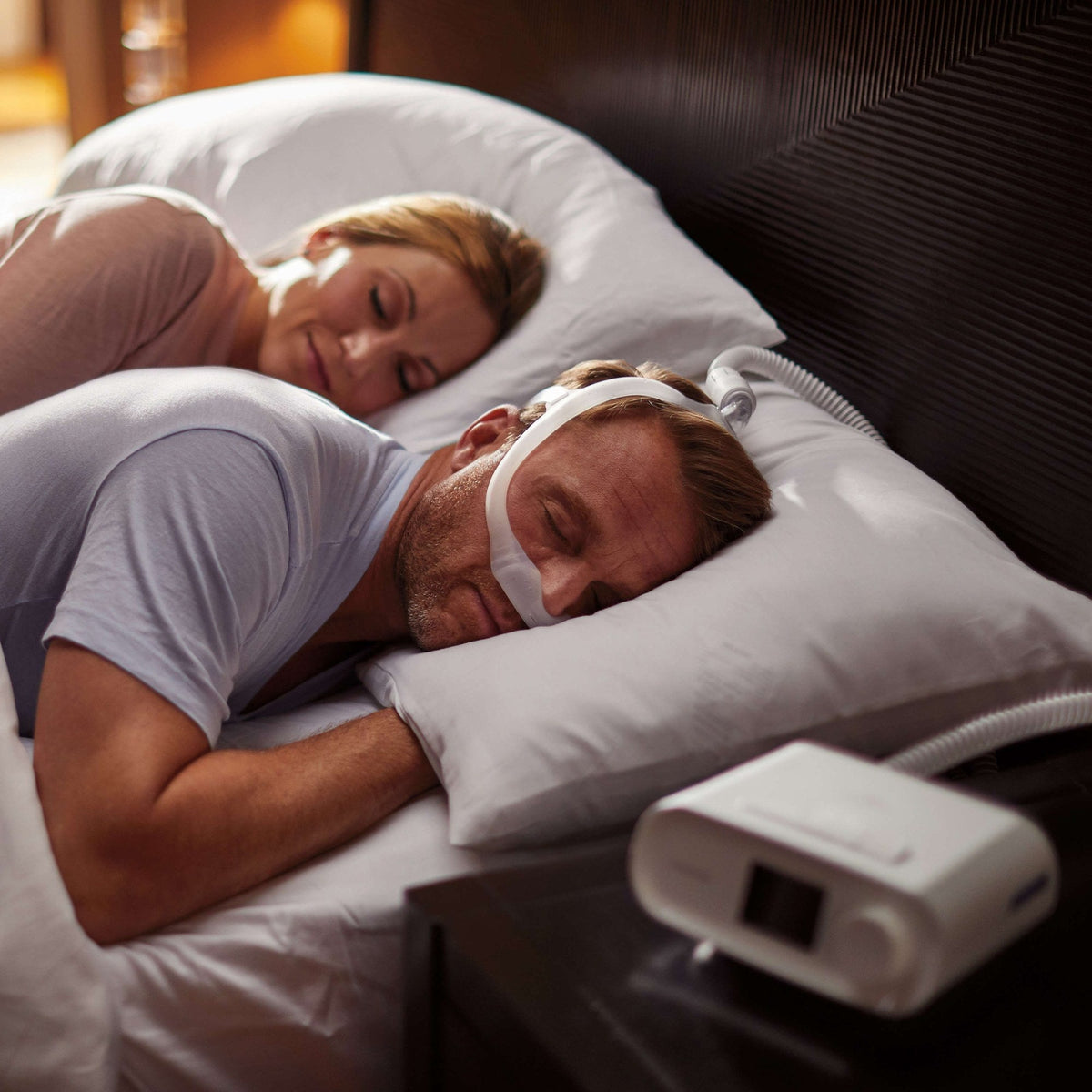 A couple asleep in bed, the man in a nasal CPAP mask, with a DreamStation on the nightstand