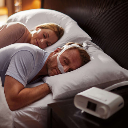 A couple asleep in bed, the man in a nasal CPAP mask, with a DreamStation on the nightstand