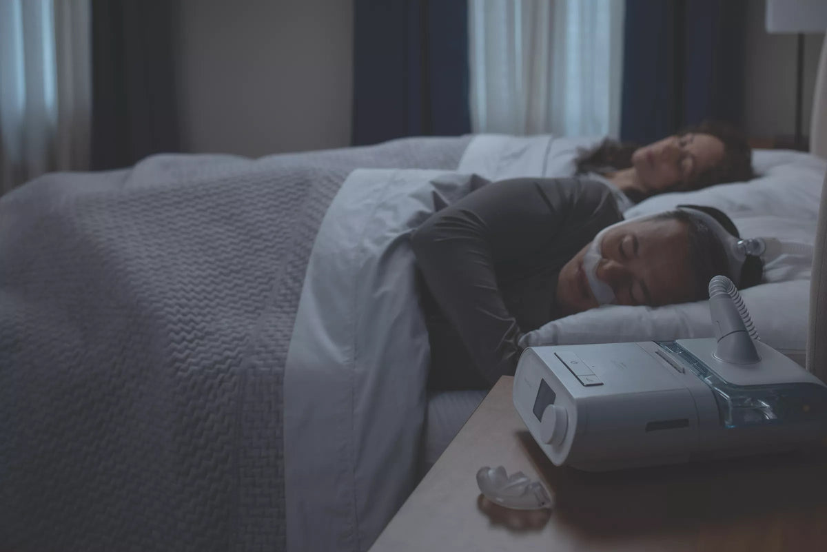 Night photo of a couple asleep in bed, one wearing the DreamWear Silicone Pillows, the machine beside them