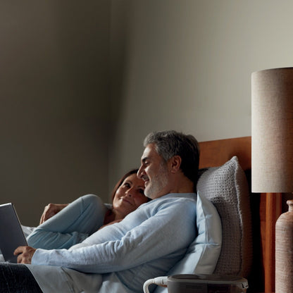 Two people relax against the headboard reading a tablet, with a DreamStation 2 and its heated tube at the bedside