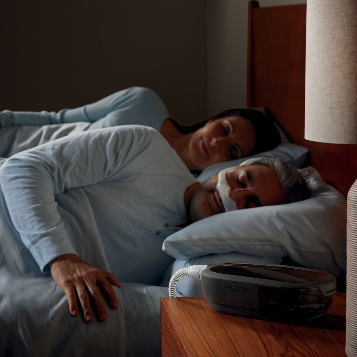 A man asleep in a nasal mask beside his partner, a DreamStation 2 and its slim tube on the nightstand