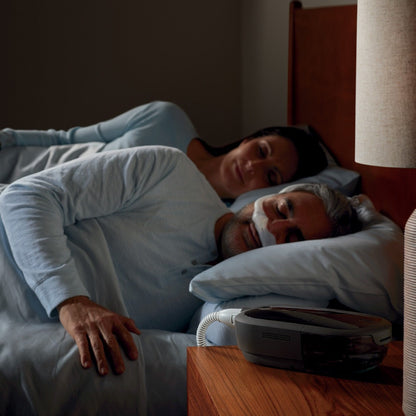 A man asleep in a nasal mask beside his partner, a DreamStation 2 and its slim tube on the nightstand