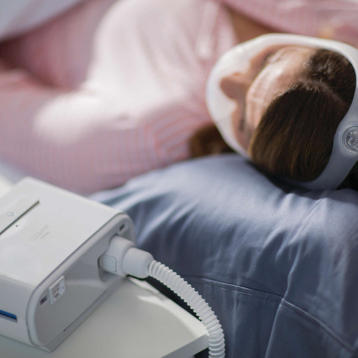 Close view of a DreamStation beside the bed with the tube running up to a sleeper's nasal mask