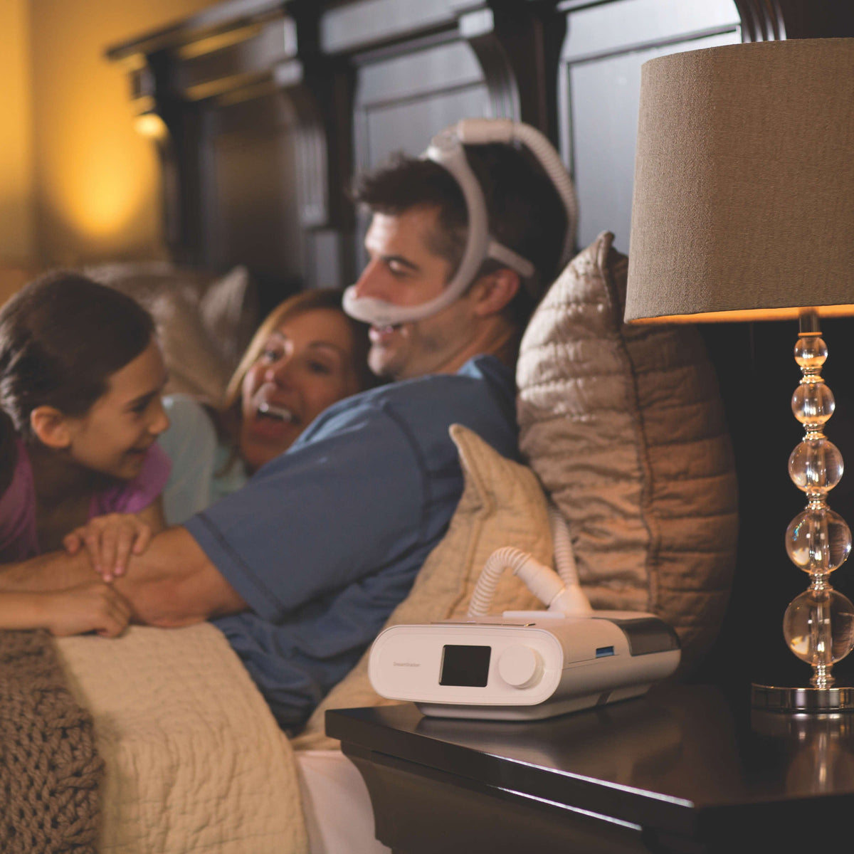 A father wearing his mask laughs with his family in bed while the DreamStation runs on the nightstand