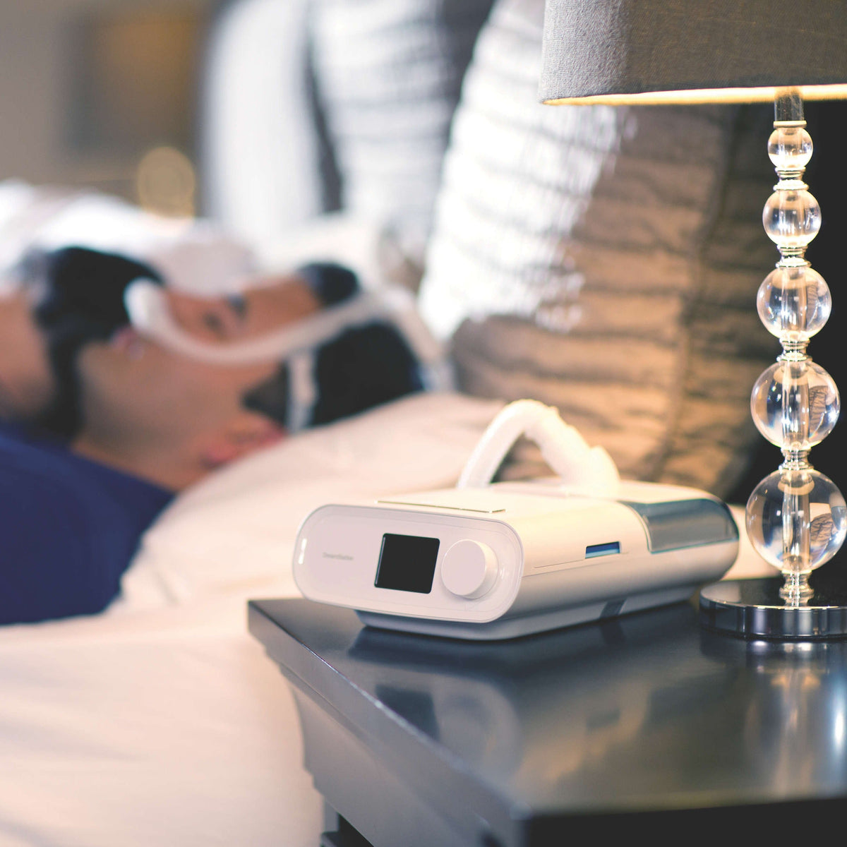 A DreamStation with its heated humidifier on a nightstand, the tube arching over toward a sleeping couple