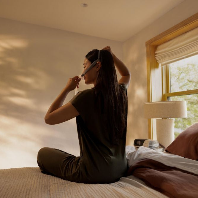 Photo of a woman sitting on her bed pulling the AirFit P10 For Her headgear over her head